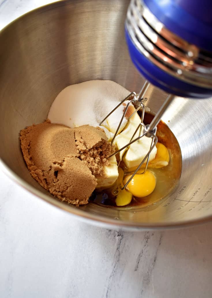 Brown sugar, white sugar, eggs, butter, and vanilla in large mixing bowl with electric hand mixer in top right corner