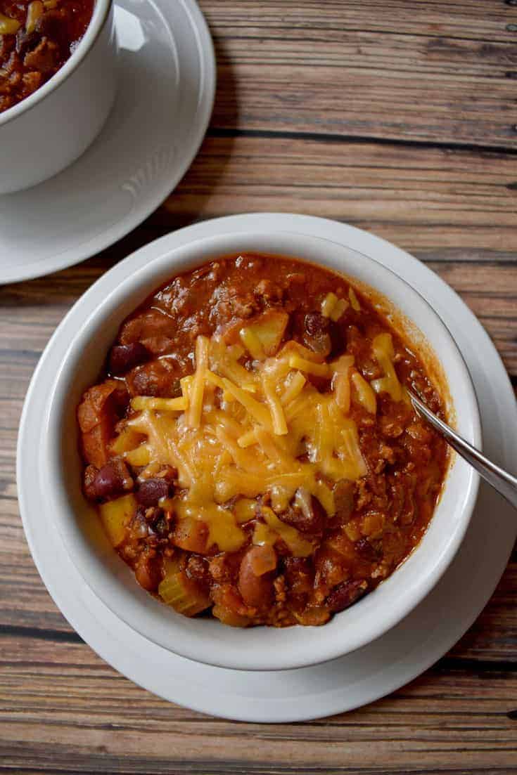 Chili with Beef, Turkey and Potatoes topped with melted cheddar cheese in white bowl on round white plate with silver spoon in bowl | Flavor Walk