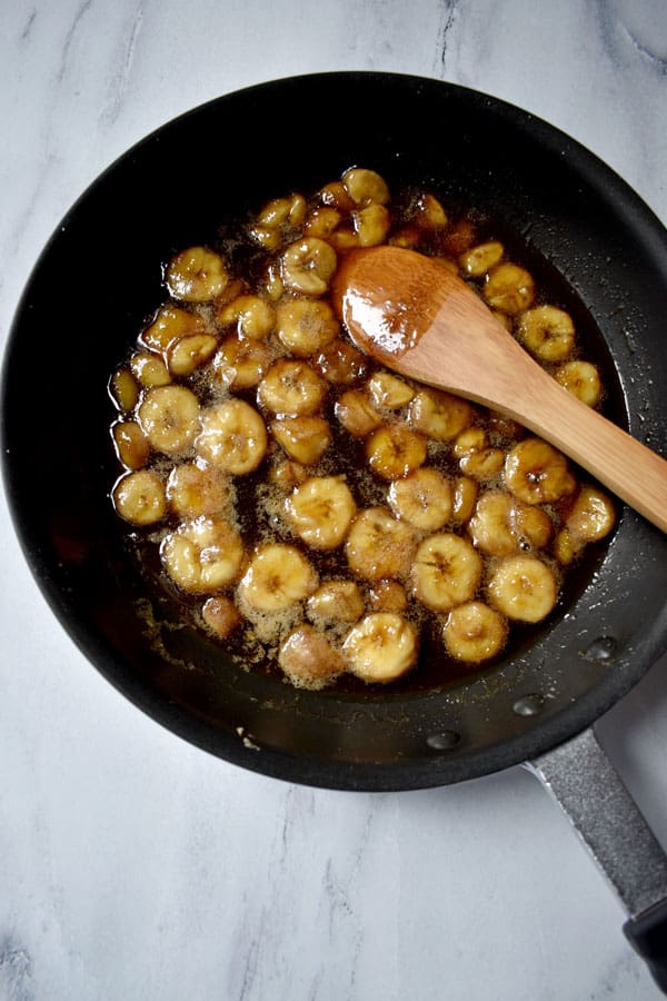 Sliced caramelized bananas in skillet with wooden spoon on granite counter top. | Flavor Walk