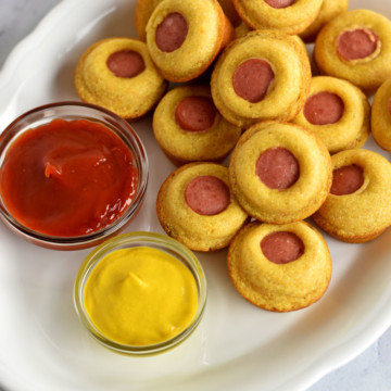 Stack of mini corn dog muffins on white plate with cups of ketchup and mustard
