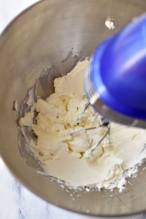 Blue electric hand mixer beating cream cheese in large mixing bowl