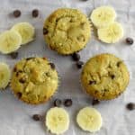 Three Gluten Free Chocolate Chip Banana Muffins on parchment paper with scattered banana slices and chocolate chips