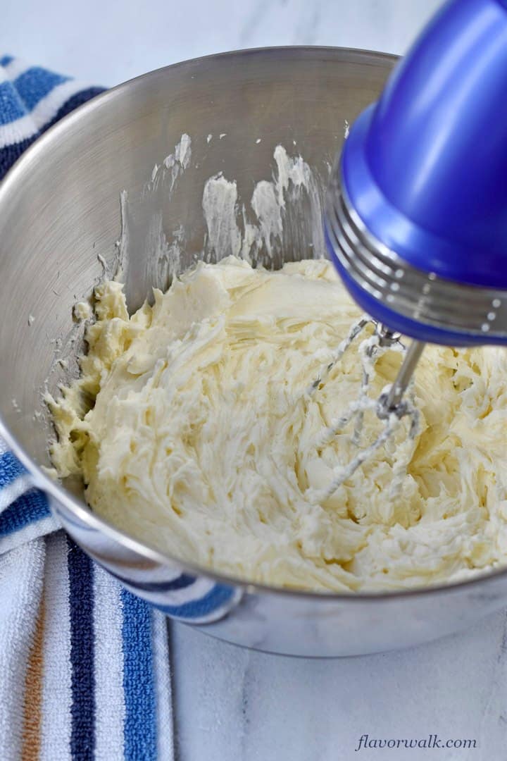 Cream cheese, sugar and vanilla beaten together in a large metal mixing bowl with blue electric hand mixer and blue and white striped kitchen towel on the left