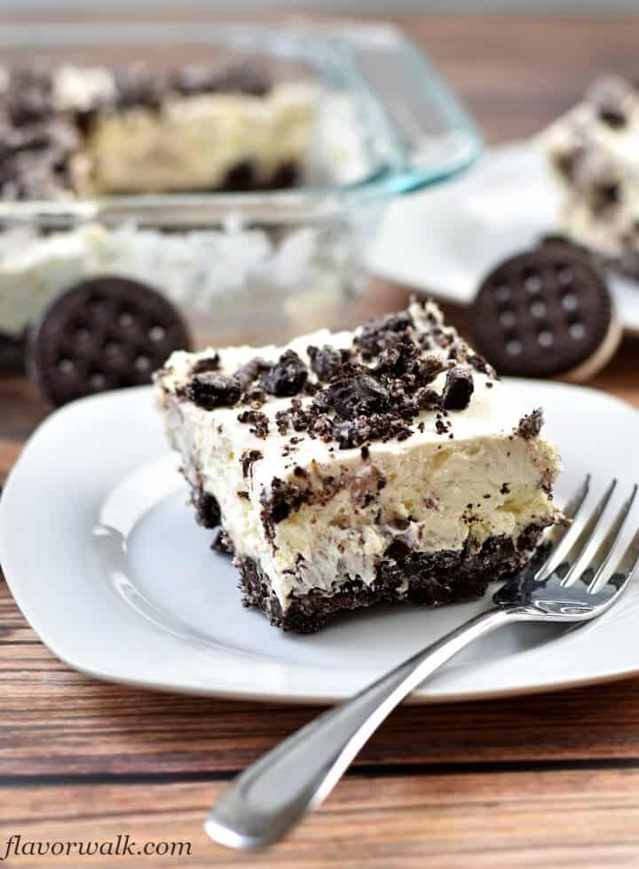 A slice of no bake oreo cheesecake and fork on white plate with a glass pan filled with remaining cheesecake in upper left corner