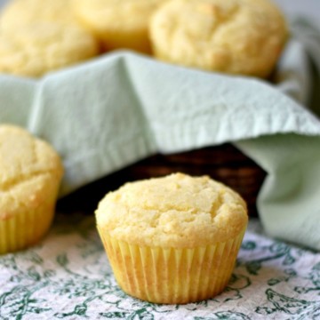 2 sweet cornbread muffins on a grean and white kitchen towel with a basket of muffins in the background