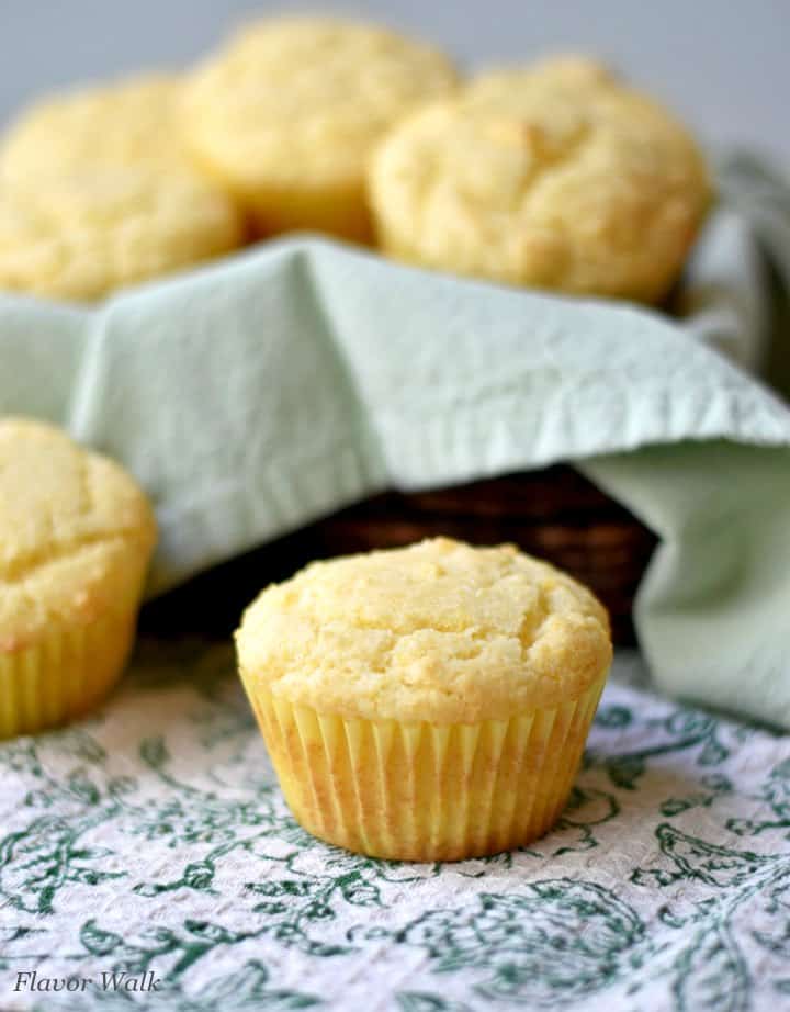 2 sweet cornbread muffins on a grean and white kitchen towel with a basket of muffins in the background