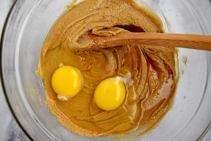 Overhead view of clear mixing bowl with peanut butter mixture, eggs, vanilla and wooden spoon