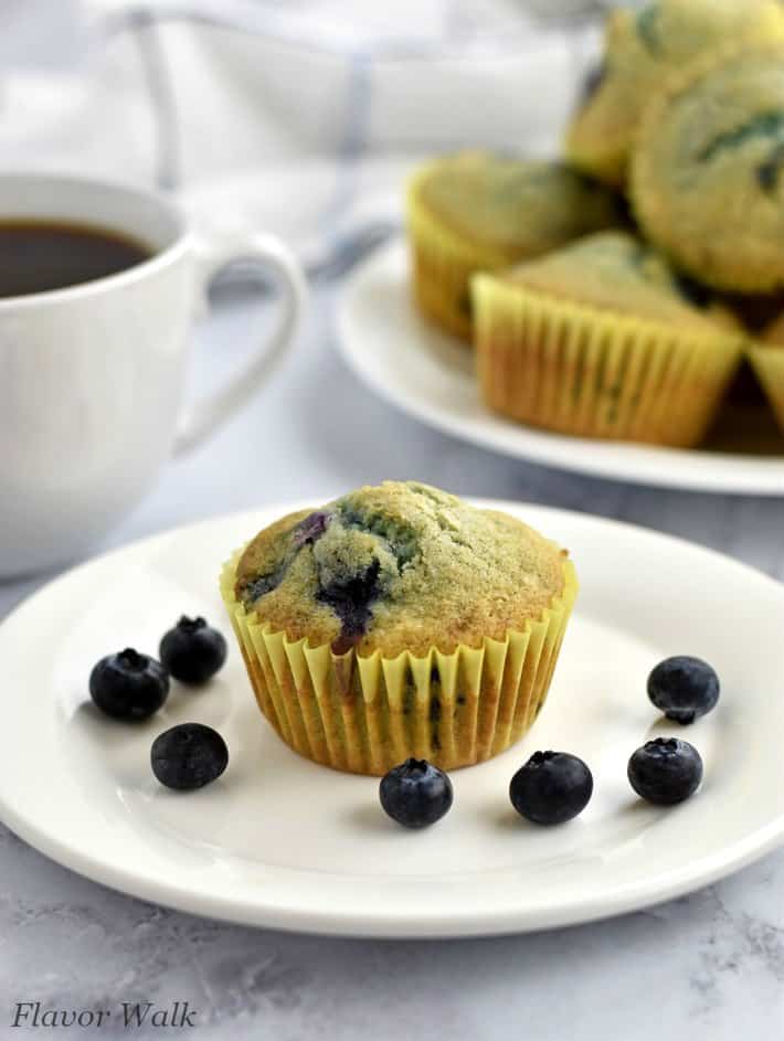 One gluten free blueberry muffin and blueberries on white plate, with more muffins in upper right corner and cup of coffee in upper left corner