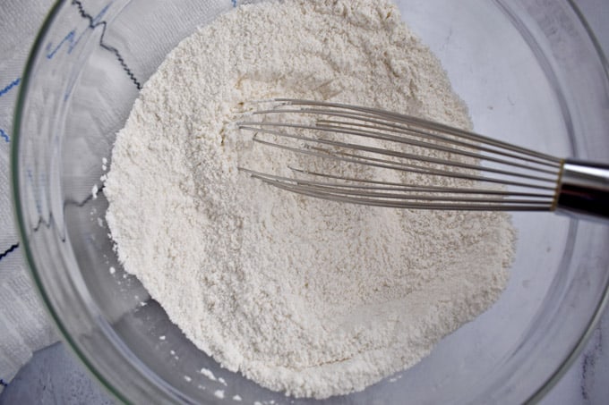 Glass mixing bowl containing a mix of gluten free flour, sugar, baking powder, and salt with wire whisk