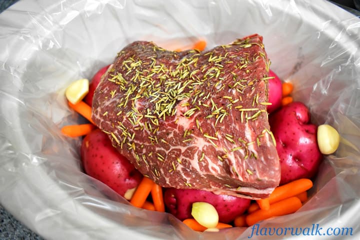 White lined slow cooker crock with uncooked beef bottom round roast on top of potatoes, carrots, and garlic cloves