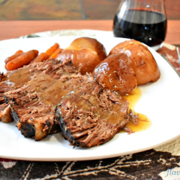 A serving of slow cooder beef pot roast and vegetables on white dinner plate with silverware to the right and a glass of red wine in the upper right corner
