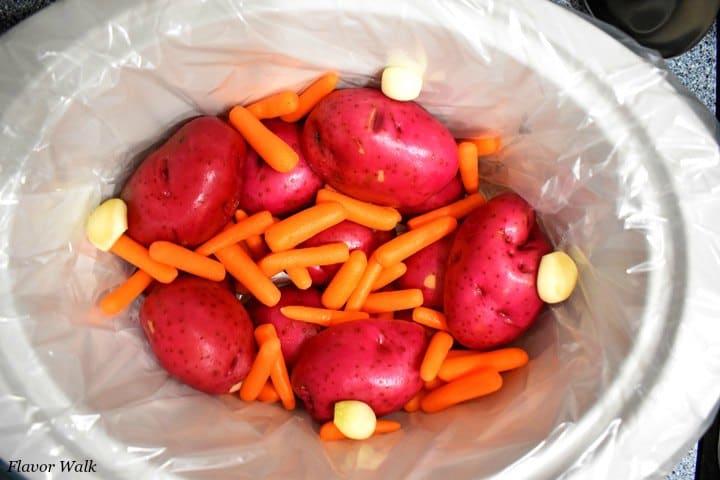 White lined slow cooker crock with red potatoes, carrots, and garlic cloves in crock