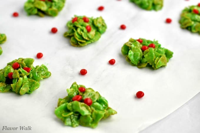 Several Holly Cookies on a sheet of parchment paper with red candies sprinkled around the cookies.