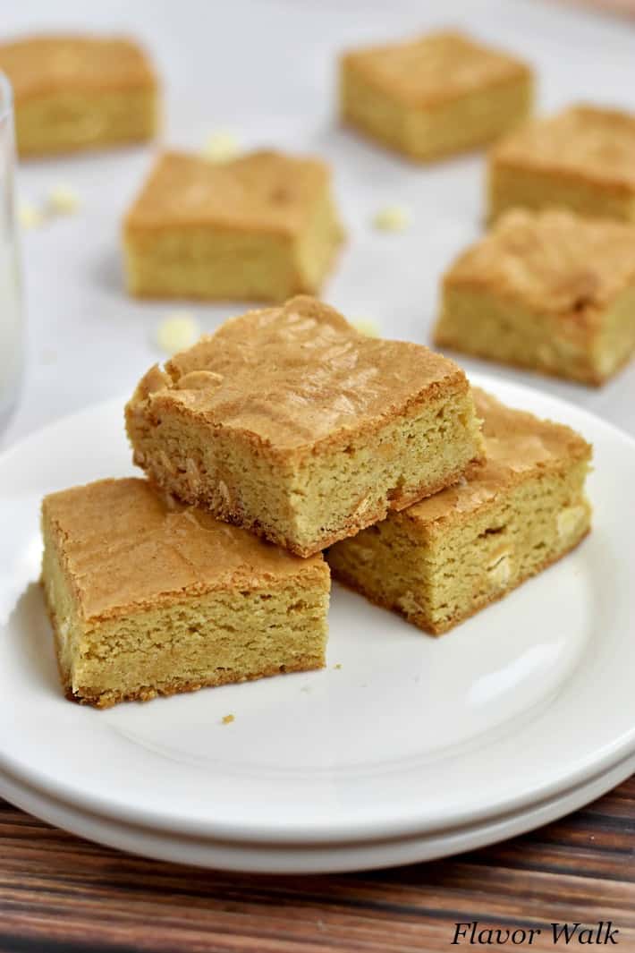 3 Gluten Free Blondies on a stack of 2 round white plates with a glass of milk and more blondies in background