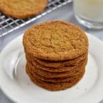Stack of gluten free ginger snaps on small white plate with glass of milk and more cookies on wire rack in the background