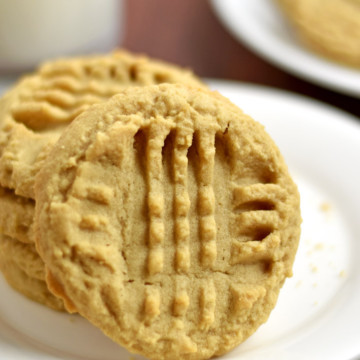 Stack of four gluten free peanut butter cookies on a small white plate with a glass of milk and more cookies in the background.