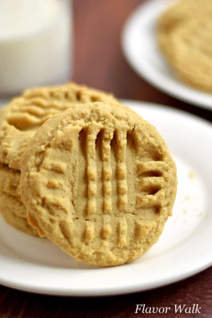 Stack of four gluten free peanut butter cookies on a small white plate with a glass of milk and more cookies in the background.