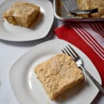 Overhead view of a piece of apple walnut crumb cake and fork on small white plate with another piece of cake and the remaining pan of cake in the background.