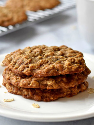 Stack of three banana oatmeal cookies on a white plate with more cookies on a wire rack in the background.