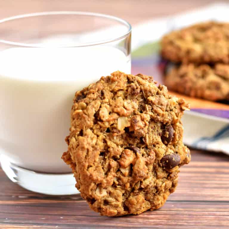 One monster cookie propped against a glass of milk with a plate of more cookies in the background.