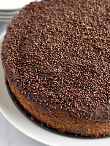 Close up view of an uncut chocolate brownie cheesecake on a round white plate with forks and smaller plates in the background.