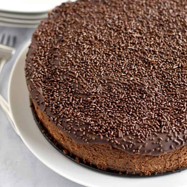 Close up view of an uncut chocolate brownie cheesecake on a round white plate with forks and smaller plates in the background.