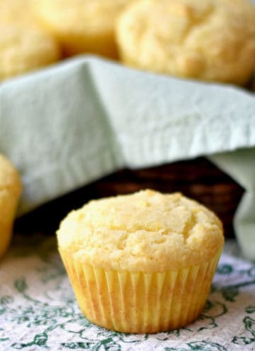 A sweet cornbread muffin on a green and white kitchen towel with a basket of muffins in the background.