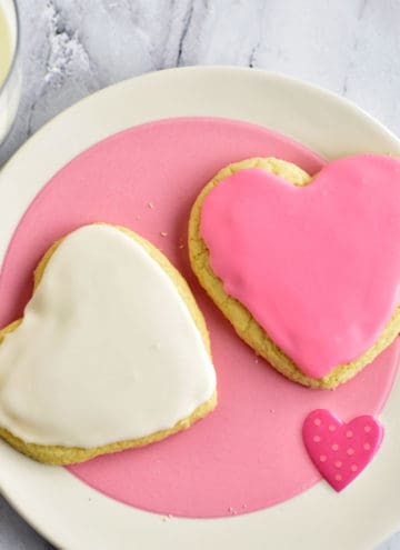 Two heart-shaped frosted sugar cookies on a pink and white plate with a glass of milk in the background.