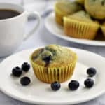 One gluten free blueberry muffin and some blueberries on a small white plate. A cup of coffee and more muffins in the background.