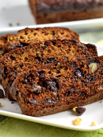 Three slices of gluten free chocolate chip banana bread and a few walnuts and chocolate chips on a square white plate with the remaining loaf in the background.