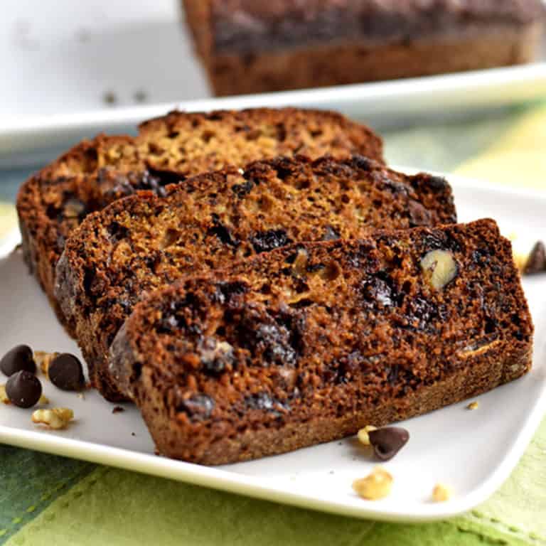 Three slices of gluten free chocolate chip banana bread and a few walnuts and chocolate chips on a square white plate with the remaining loaf in the background.