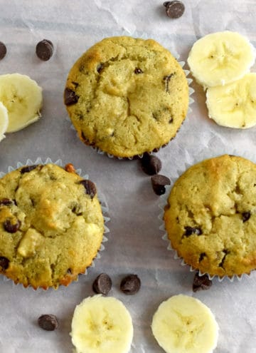Overhead view of three chocolate chip banana muffins on parchment paper with sliced bananas and chocolate chips scattered around the muffins.