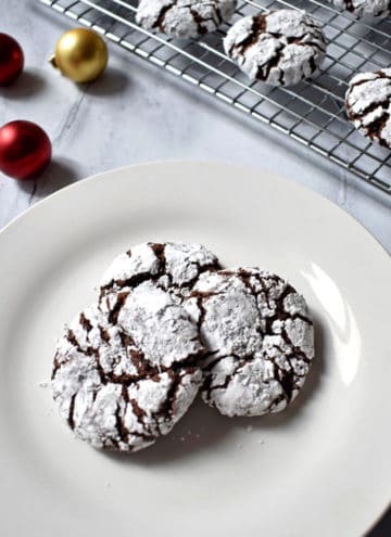 Three gluten free chocolate crinkle cookies on a small white plate with two small red ornaments and one gold ornament in the upper left corner and more cookies on a rack in the upper right corner.
