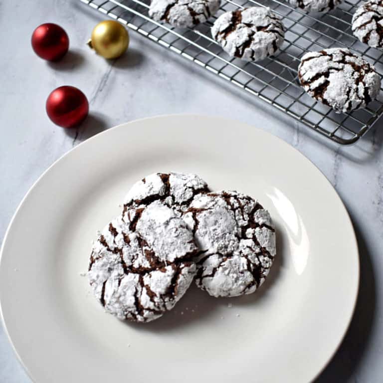 Three gluten free chocolate crinkle cookies on a small white plate with two small red ornaments and one gold ornament in the upper left corner and more cookies on a rack in the upper right corner.