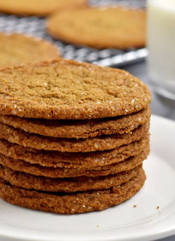 A stack of gluten free ginger snaps on a small white plate with more cookies on a wire rack and a glass of milk in the background.