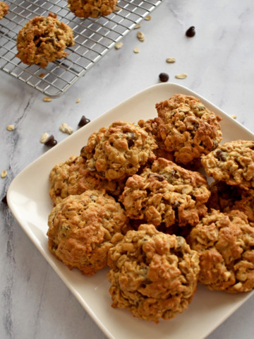 A square white plate filled with gluten free oatmeal cookies with a sprinkling of chocolate chips and oats and more cookies in the background.