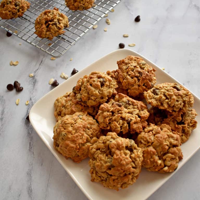 A square white plate filled with gluten free oatmeal cookies with a sprinkling of chocolate chips and oats and more cookies in the background.