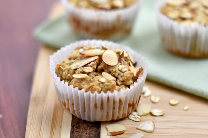Close up view of a gluten free oatmeal muffin and a few oats and almond slices on a wood cutting board with additional muffins in the background.