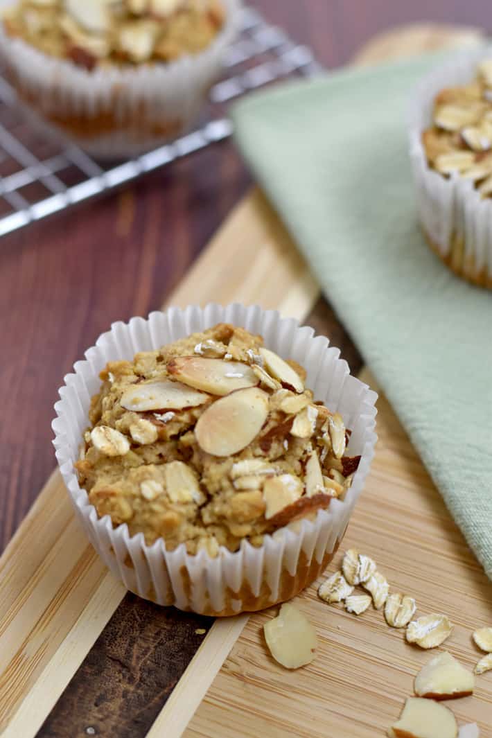 A gluten free oatmeal muffin and a few oats and slivered almonds on a wood cutting board with additional muffins in the background.