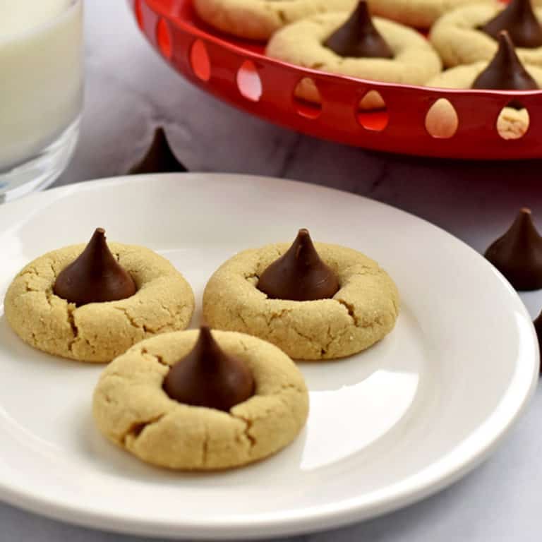 Three gluten free peanut butter blossoms on a small white plate with unwrapped Hershey's kisses, a glass of milk, and more blossoms in the background.