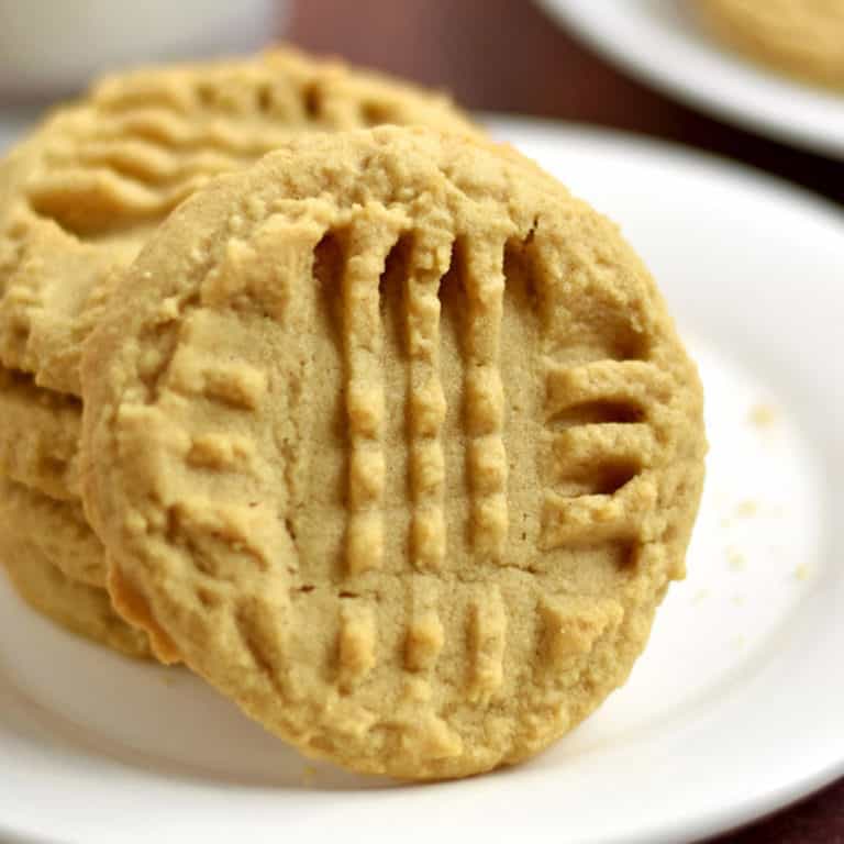 Close up of a gluten free peanut butter cookie propped against a stack of three more cookies on a white plate.