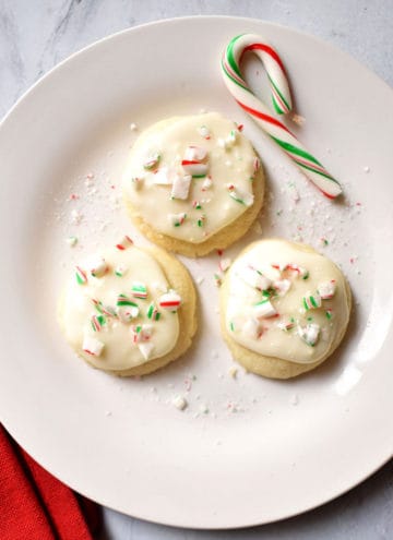 Three peppermint meltaway cookies and a small candy cane on a small white plate with a red kitchen towel on the left of the plate.