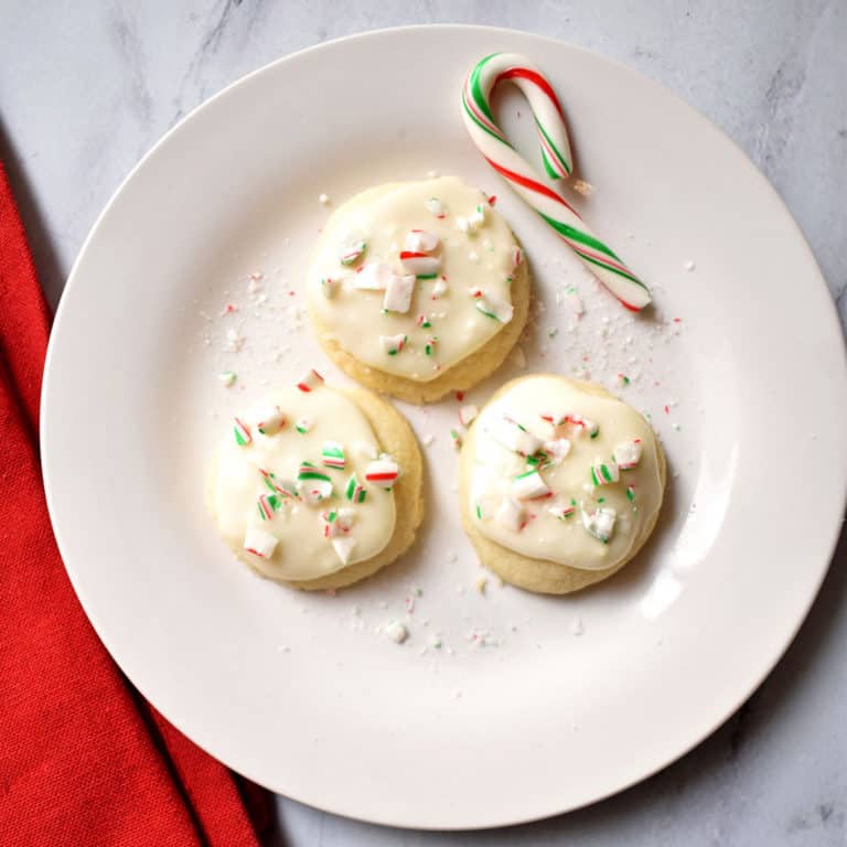 Three peppermint meltaway cookies and a small candy cane on a small white plate with a red kitchen towel on the left of the plate.