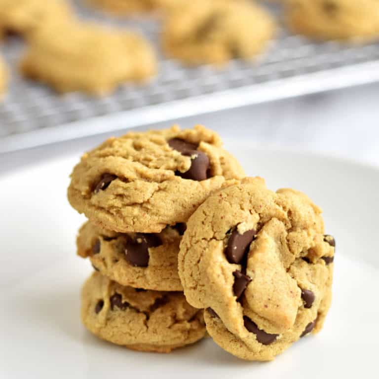 One chocolate chip almond butter cookie propped against a stack of three cookies on a white plate with more cookies in the background.