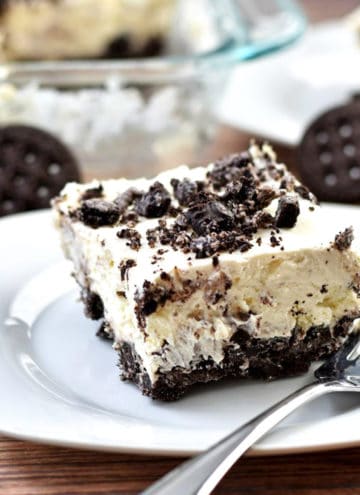 A serving of no bake oreo cheesecake and a fork on a white plate with additional cheesecake in the background.