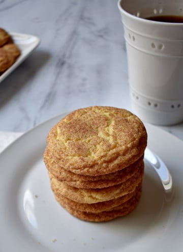 A stack of old-fashioned snickerdoodle cookies on a white plate with a cup of coffee and more cookies in the background.