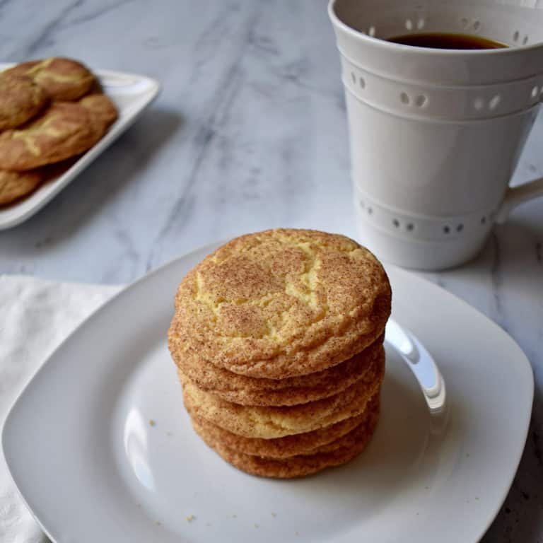 A stack of old-fashioned snickerdoodle cookies on a white plate with a cup of coffee and more cookies in the background.