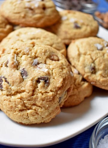 Close up view of a white plate filled with salted caramel chocolate chip cookies with a small container of sea salt in the lower right corner/