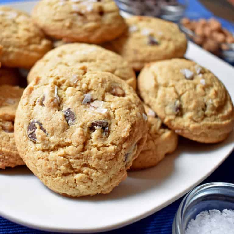 Close up view of a white plate filled with salted caramel chocolate chip cookies with a small container of sea salt in the lower right corner/