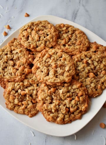 Overhead view of a square white plate filled with butterscotch oatmeal cookies with butterscotch chips and shredded coconut scattered around the plate.